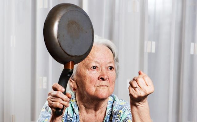 An elderly woman holding up a pan, looking like she is threatening somebody