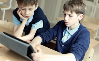 Two young boys wearing light-blue shirts and navy sweaters as school uniforms and one is using a tab...
