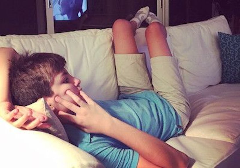 A teenage boy wearing a blue T-shirt and beige shorts while lying on a couch and watching TV.