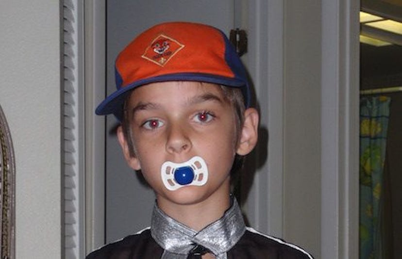 A quirky kid with a red cap on his head holding a baby pacifier in his mouth