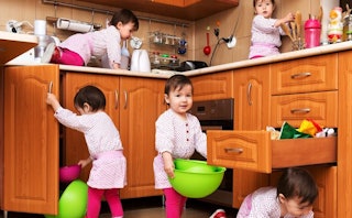 A baby in a pink playsuit in several places around the kitchen floor and counter