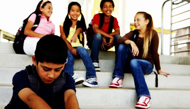 A bullied boy sitting alone on the stairs while four schoolmates are laughing at him.