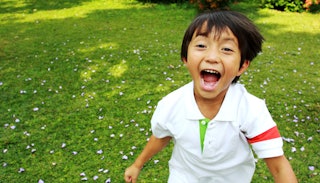 A small kid running, screaming, and laughing in a white shirt on a grass field