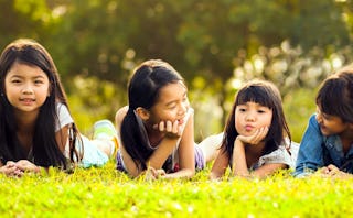 Four long-haired brunette girls lying on their stomachs on bright green grass, looking to the side a...