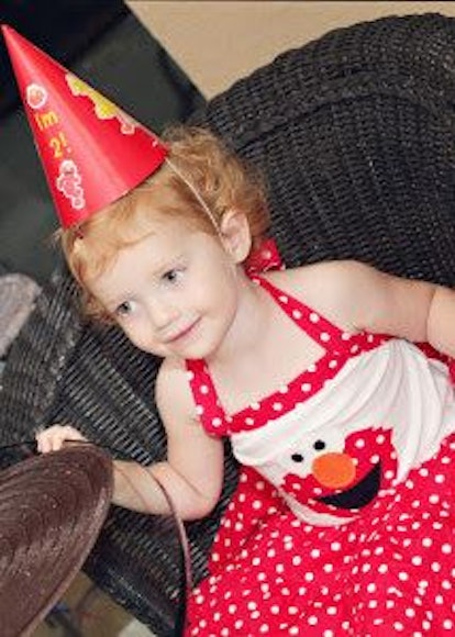 A little blonde girl wearing a red and white Elmo dress with polka dots and a red party hat with "I'...