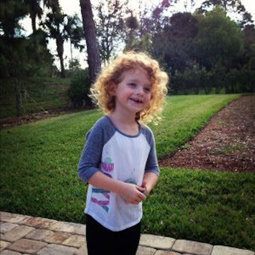 A little blonde girl wearing a white and gray long sleeve t-shirt while standing in a backyard and s...