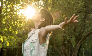 A woman in a blue denim vest with her arms raised standing in a forest