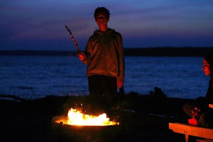 A woman holding a wooden stick near a campfire, while her child sits on the side 
