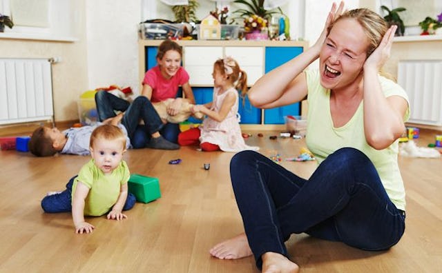 A mother screaming on the floor with her children making a mess in the background while her spouse i...