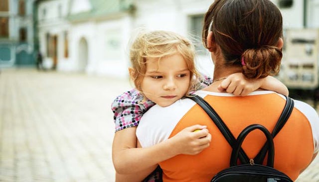 A mom in an orange shirt with a backpack carrying her child while walking on the street