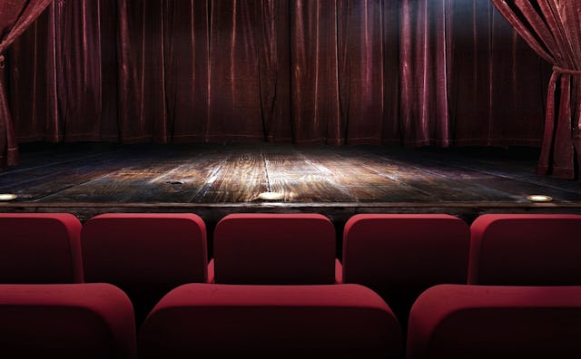 A dark-brown wooden stage with lights pointed at the center part, a red plush curtain, and red seats...