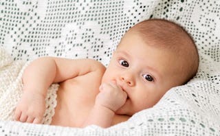 A baby lying on a white couch while holding his fingers inside his mouth whose parents have to make ...