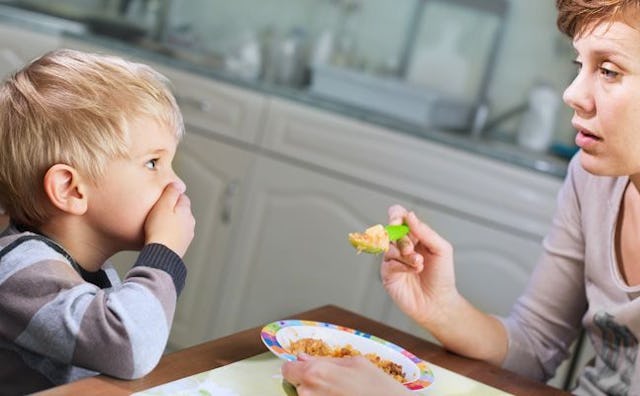 A mother trying to feed her blonde boy in a gray-blue shirt, who is refusing food by covering his mo...