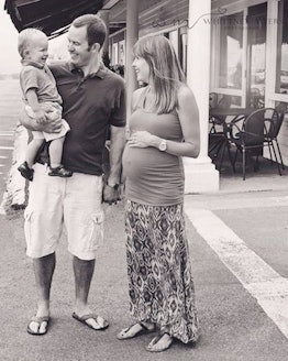 A pregnant wife standing next to her husband holding their toddler on a summer day