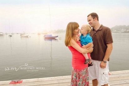 A pregnant woman and her husband standing in front of a marina, holding their toddler and smiling