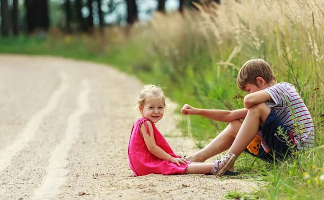 A boy sitting on the grass at the side of the road, and a girl in a pink dress sitting in front of h...
