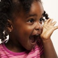 A toddler in a pink shirt with her hand next to her face with an excited facial expression