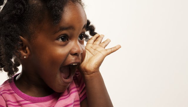 A toddler in a pink shirt with her hand next to her face with an excited facial expression