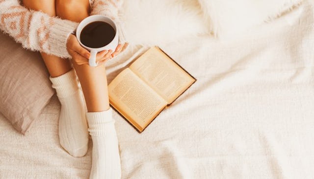 A homebody mom sitting on the bed in a comfy sweater and socks next to a book, holding a cup of coff...
