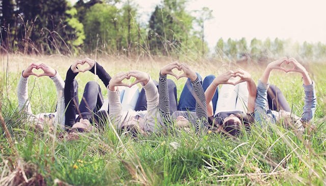 Six teenage girls lying down in a grass field with their arms raised showing heart signs formed with...