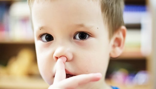 A brown-haired boy with brown eyes looking straight and holding his finger next to his nose
