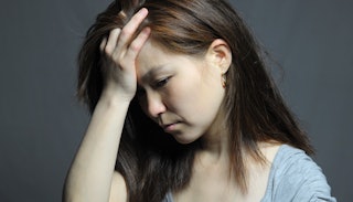 A woman with brown hair wearing a blue shirt while holding her hand on her face and looking worried 