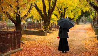 A woman in black with an umbrella walking through a leafy park during fall 