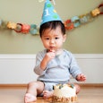 A toddler in a light blue shirt and shorts and a birthday hat sitting on the floor and eating cake f...