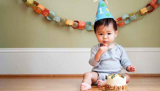 A toddler in a light blue shirt and shorts and a birthday hat sitting on the floor and eating cake f...