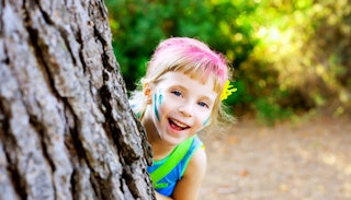 A smiling 4-year-old peeking from behind a tree with hair dyed pink
