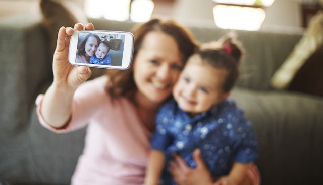 A single mom taking a selfie with her daughter using a smartphone back camera while smiling and sitt...