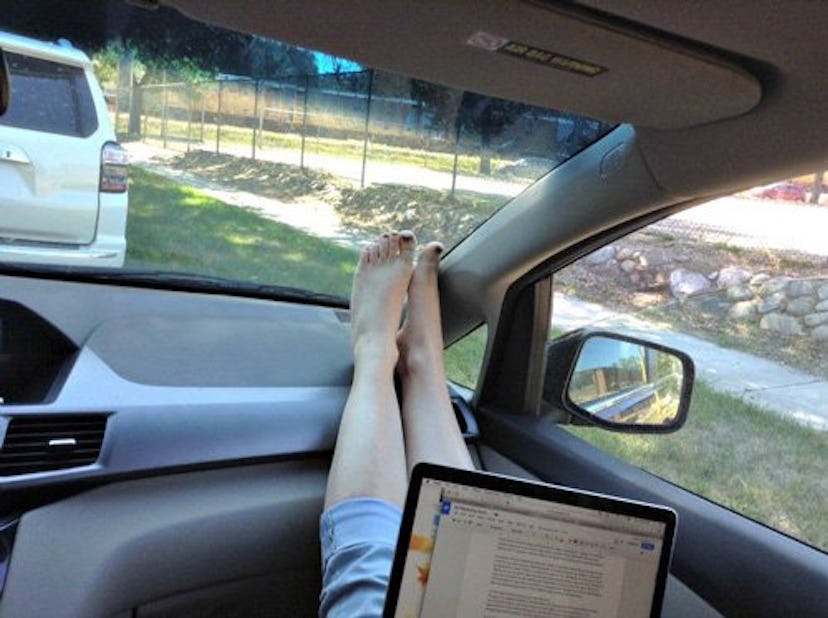 A woman with her feet up on the dashboard of a minivan, working on a laptop.