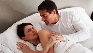 A man in a white shirt with an manner lying next to woman in bed who looks at him with disapproval w...