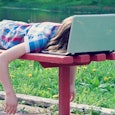 A teenager wearing a blue and red striped shirt and blue shorts sleeping on a bench with her head on...