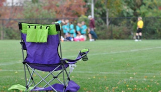 A  soccer mom's green-blue folding chair on a soccer field, with kids blurred in the distance