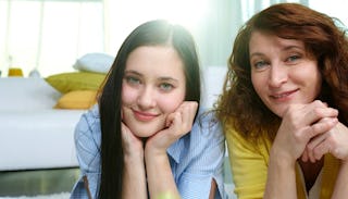 A mother lying stomach down, posing and smiling with her teenage daughter in a bedroom