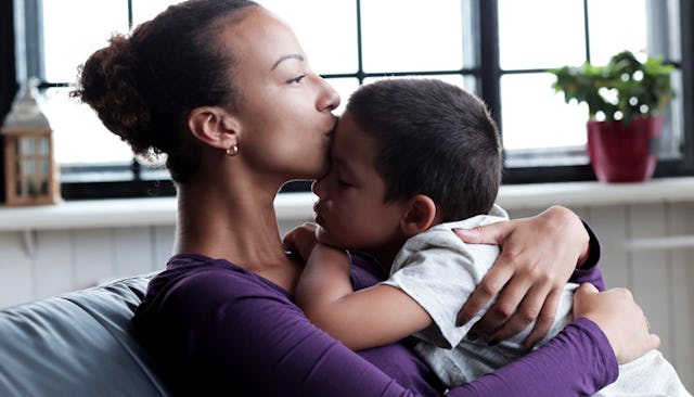 A mother with her hair up in a purple shirt and small hoop earrings sitting, hugging and kissing her...