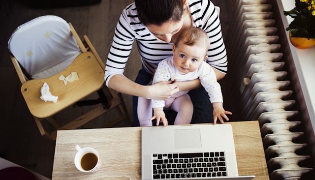 A mommy blogger sitting and holding her baby with a laptop and a cup of coffee on a desk in front of...