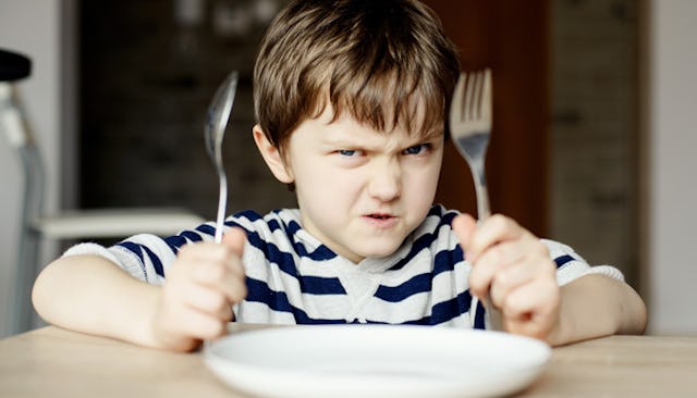 An angry boy in a navy and white striped shirt waiting for the dinner holding a fork and a knife