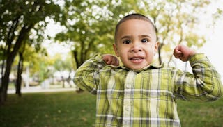 A dark-haired boy in a yellow and white checked shirt outside with both of his arms up