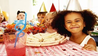 Five kids at a child’s birthday party wearing birthday hats while smiling and eating at the table