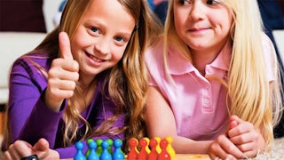 Two little girls ready to play a board game smiling and happy and one of them is holding her thumb u...