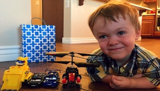 A boy with dwarfism is smiling on the floor next to his toy helicopter, excavator, and cars.