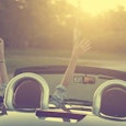 A person driving in a silver cabriolet with their hands in the air during a sunset, leaving their ki...