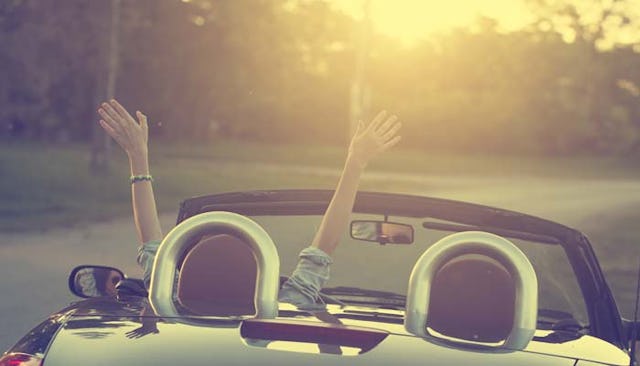 A person driving in a silver cabriolet with their hands in the air during a sunset