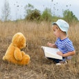 A boy in a bucket hat and a striped shirt reading a book to his teddy bear in a field