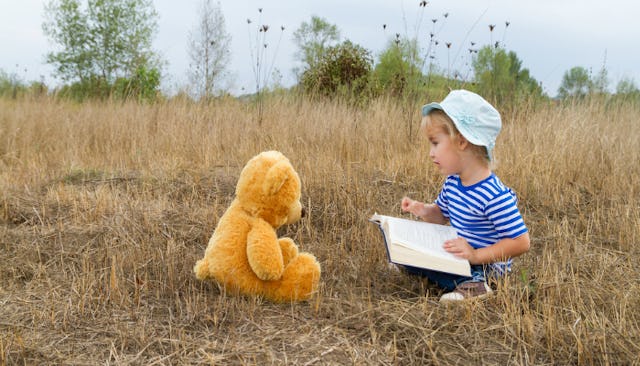 A boy in a bucket hat and a striped shirt reading a book to his teddy bear in a field