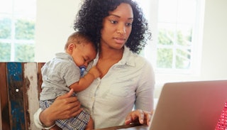 A woman holding her baby and scrolling through Facebook on her laptop 