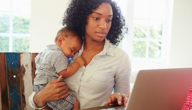 A woman holding her baby and scrolling through Facebook on her laptop