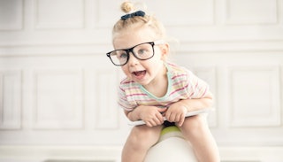 A little girl sitting on the potty, wearing a bun, glasses and a multi-colored stripe shirt